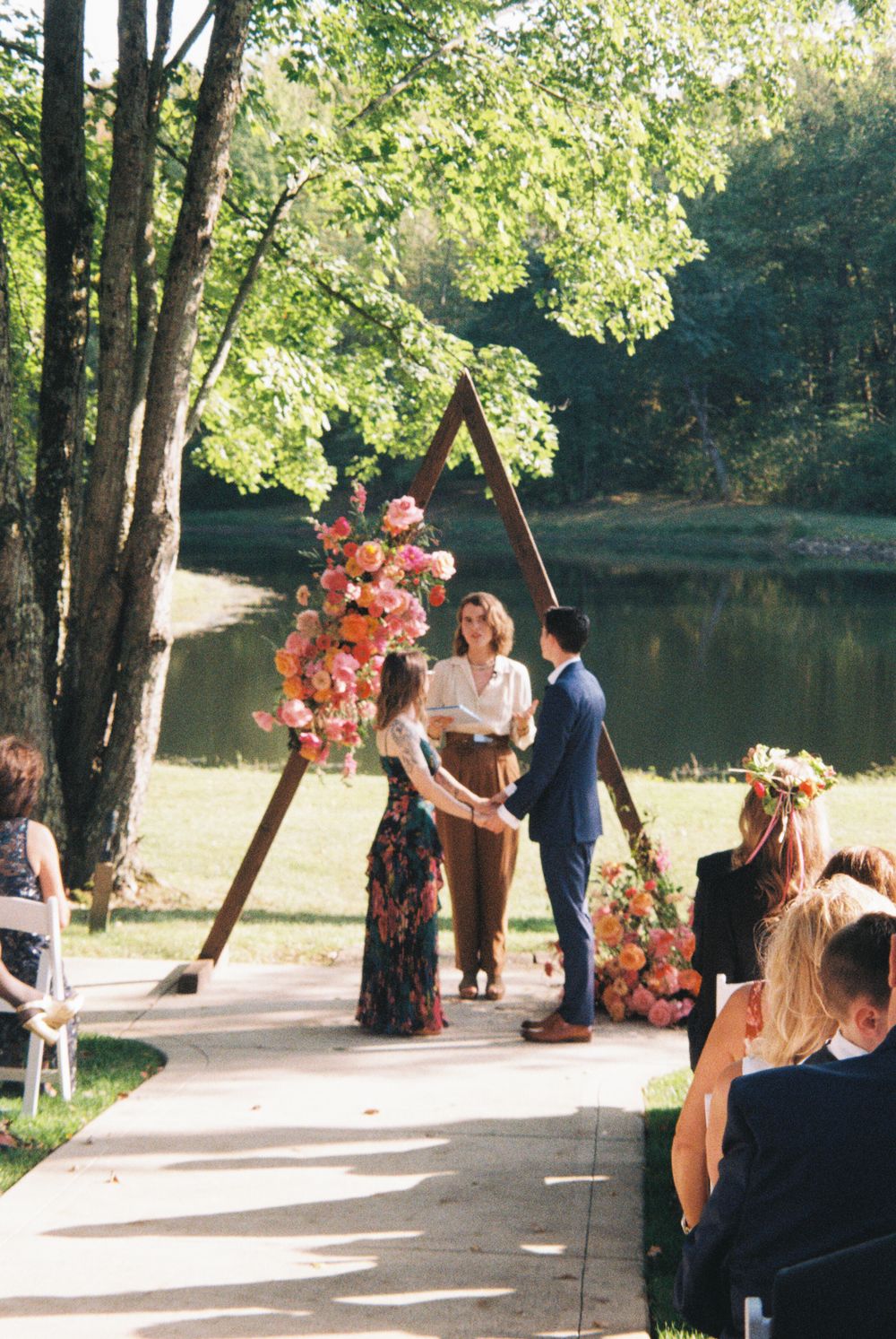 Outdoor wedding ceremony by a pond with a triangular wooden arch adorned with pink and orange flowers.