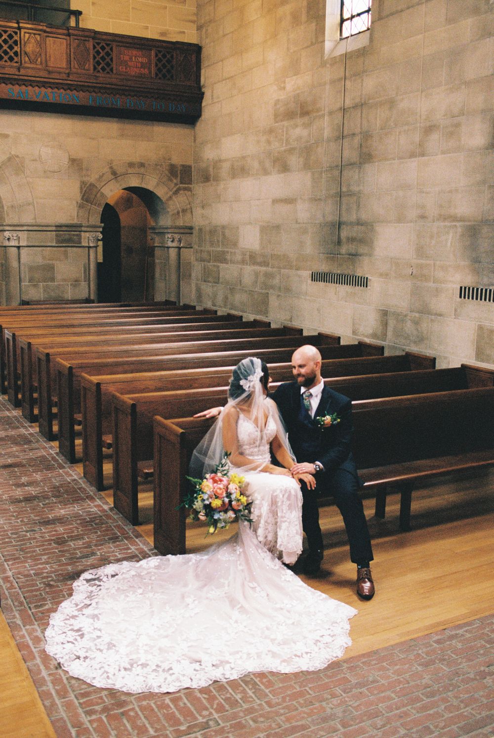 Bride and groom sit together on wooden church pews, bride wearing lace gown with veil holding colorful bouquet.