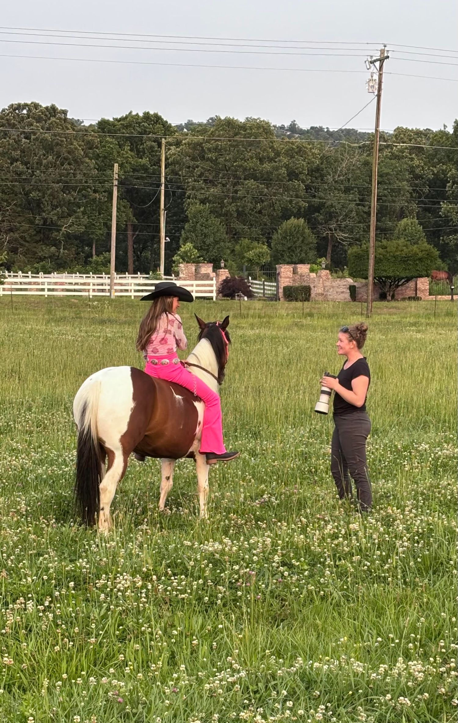 Photographer sitting in grass capturing woman in white dress posing with brown horse in green meadow near wooden fence.