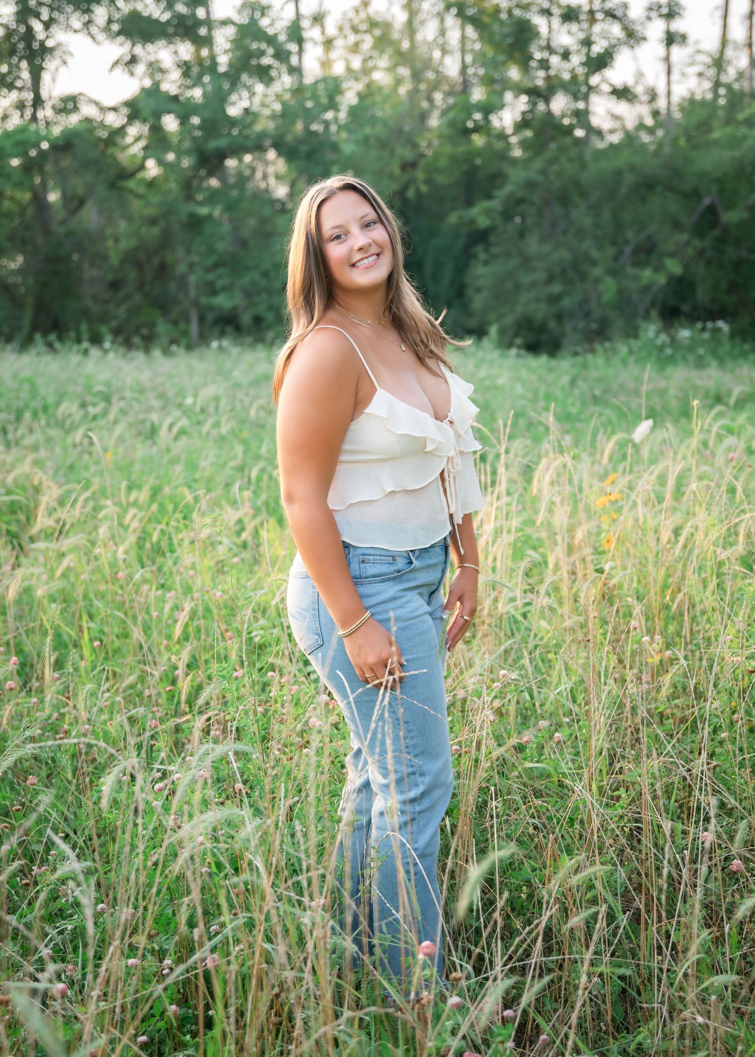 Young woman in white ruffled top and jeans smiling in a lush green meadow during golden hour.