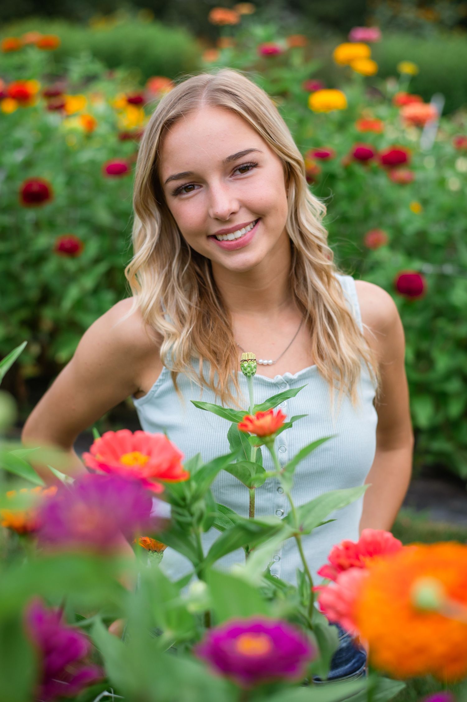 Smiling young blonde woman posing among colorful zinnia flowers in a vibrant garden setting.