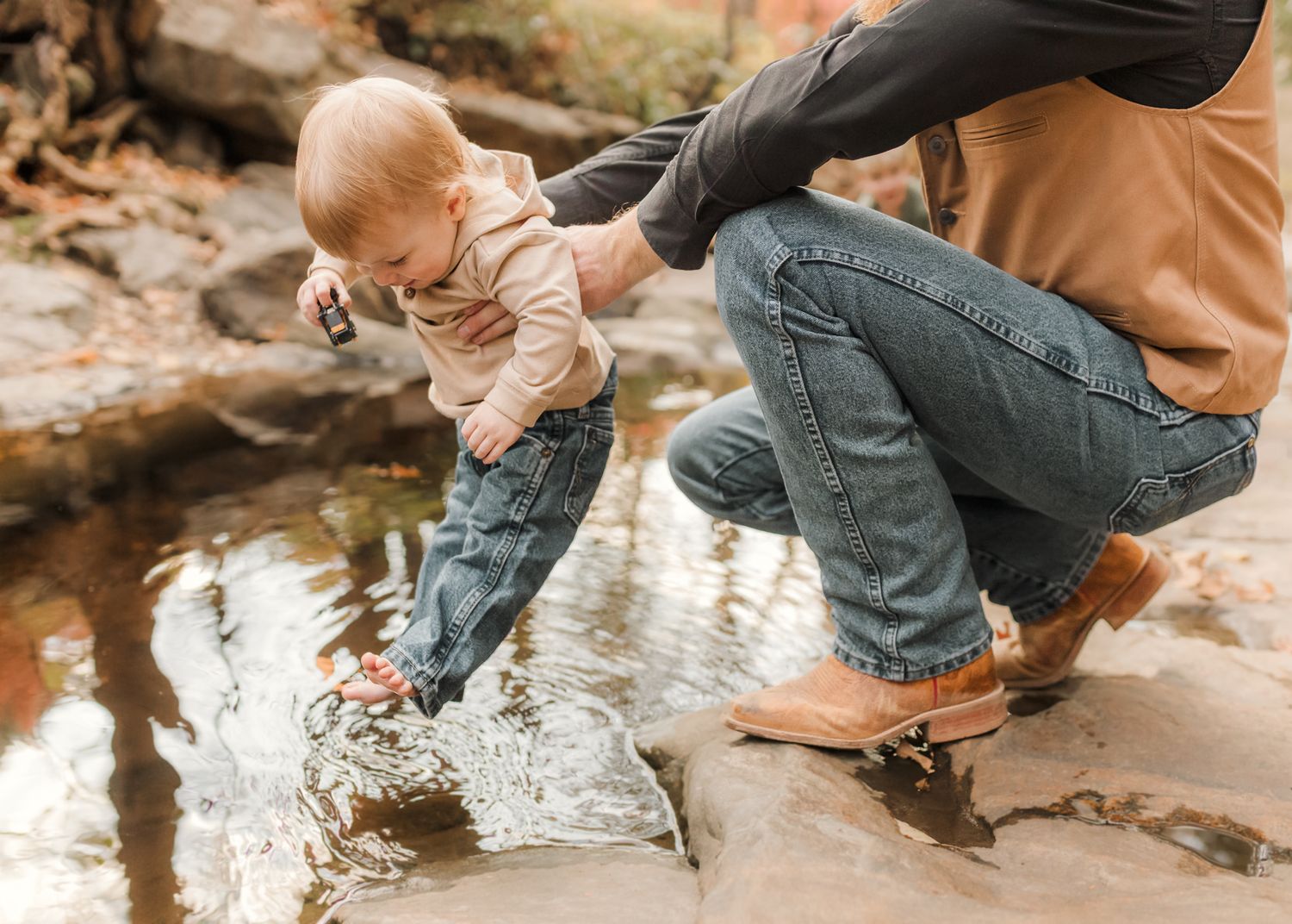 Adult helping a curious toddler touch water in a rocky stream outdoors on a fall day.