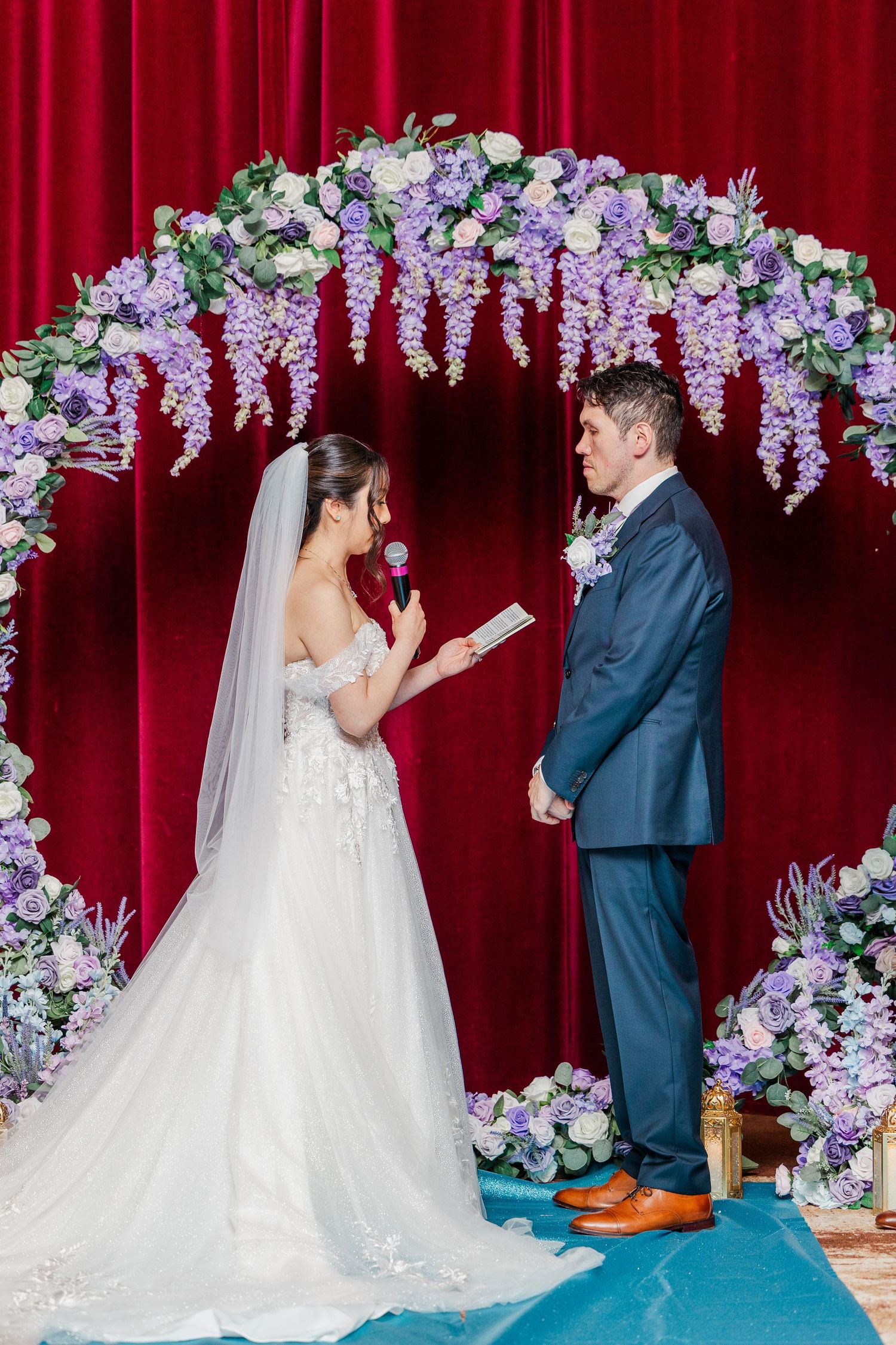 Bride reads vows to groom under floral arch with purple flowers and red velvet backdrop at elegant wedding ceremony.