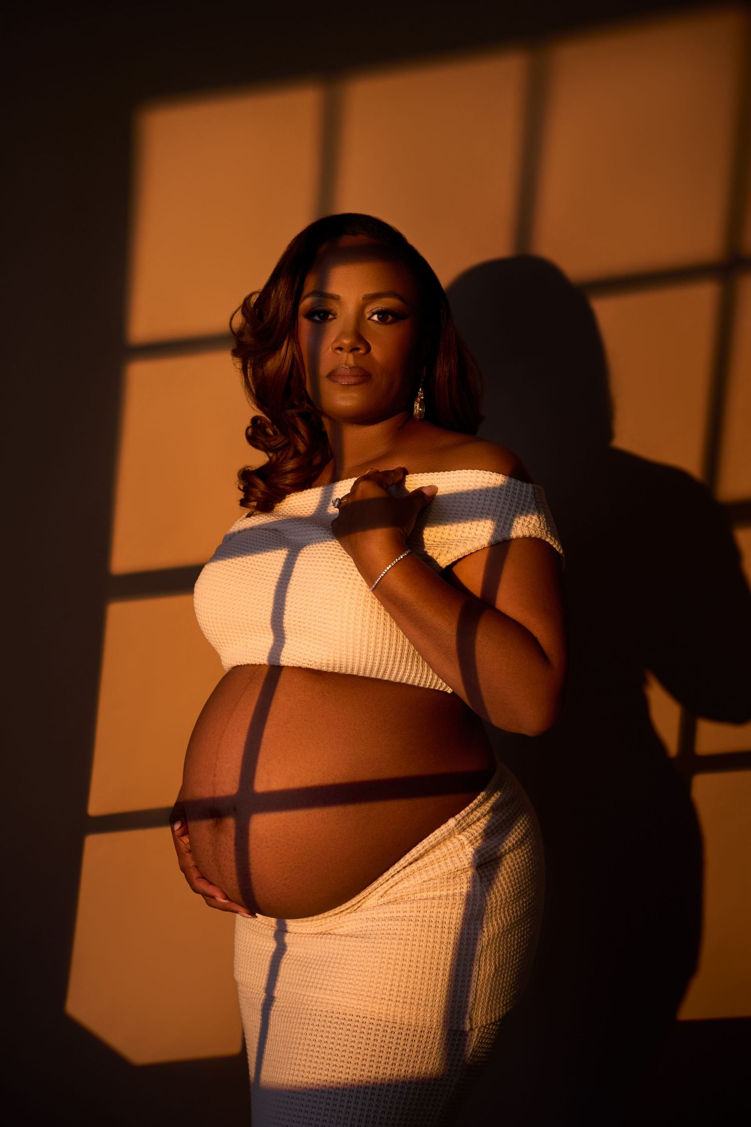 Pregnant Black woman in white crop top poses gracefully with warm golden light casting window shadows on her bare belly. NJ Maternity Photog