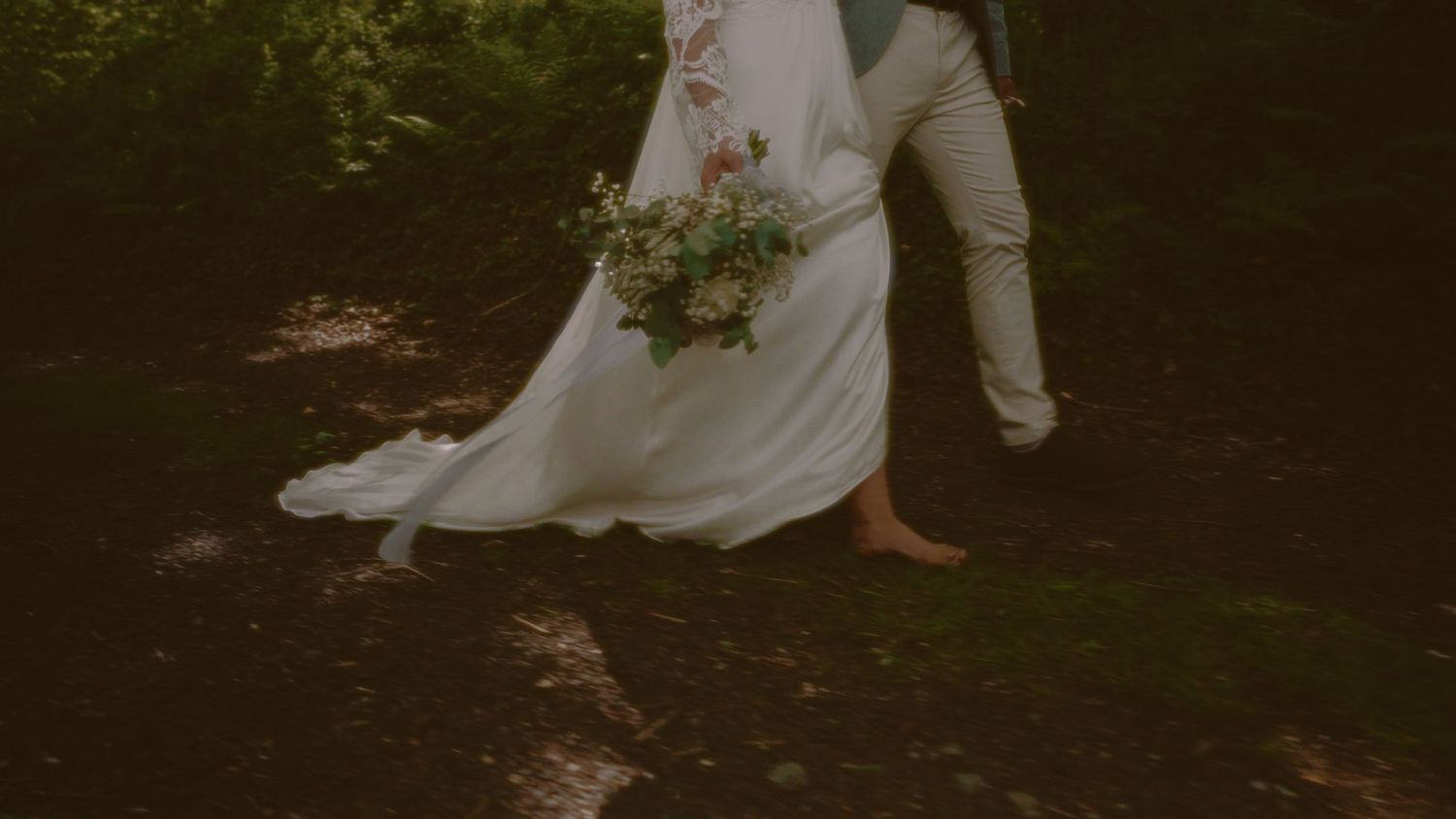 Bride and groom walking barefoot on a forest path, bride holding a wildflower bouquet in flowing white gown.
