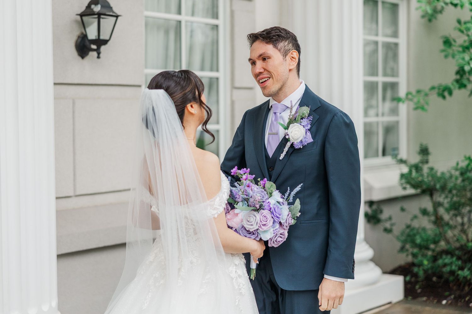 Bride in white gown with veil holds purple bouquet facing groom in navy suit at elegant white columned venue.