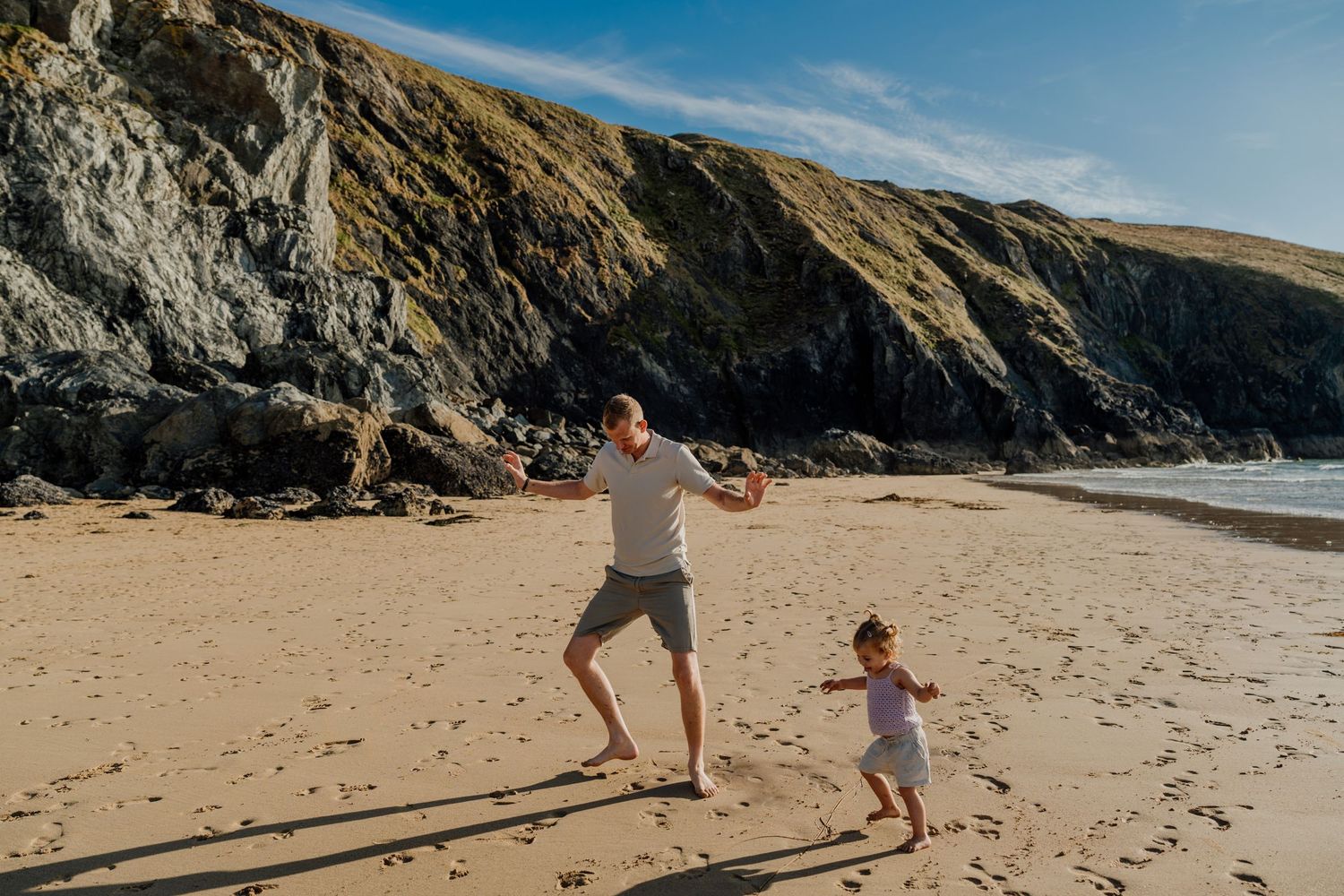 Father and young child playing and running together on a sandy beach with rocky cliffs in the background.