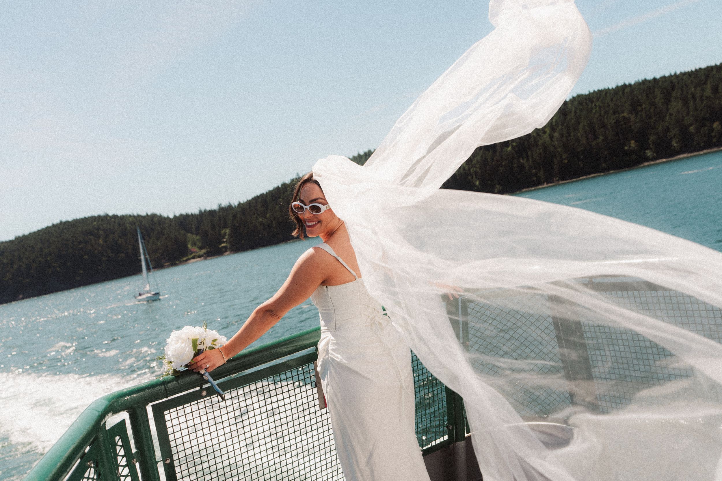 Bride in a white gown and sunglasses holds a bouquet on a ferry deck as her long veil blows in the wind over the water.