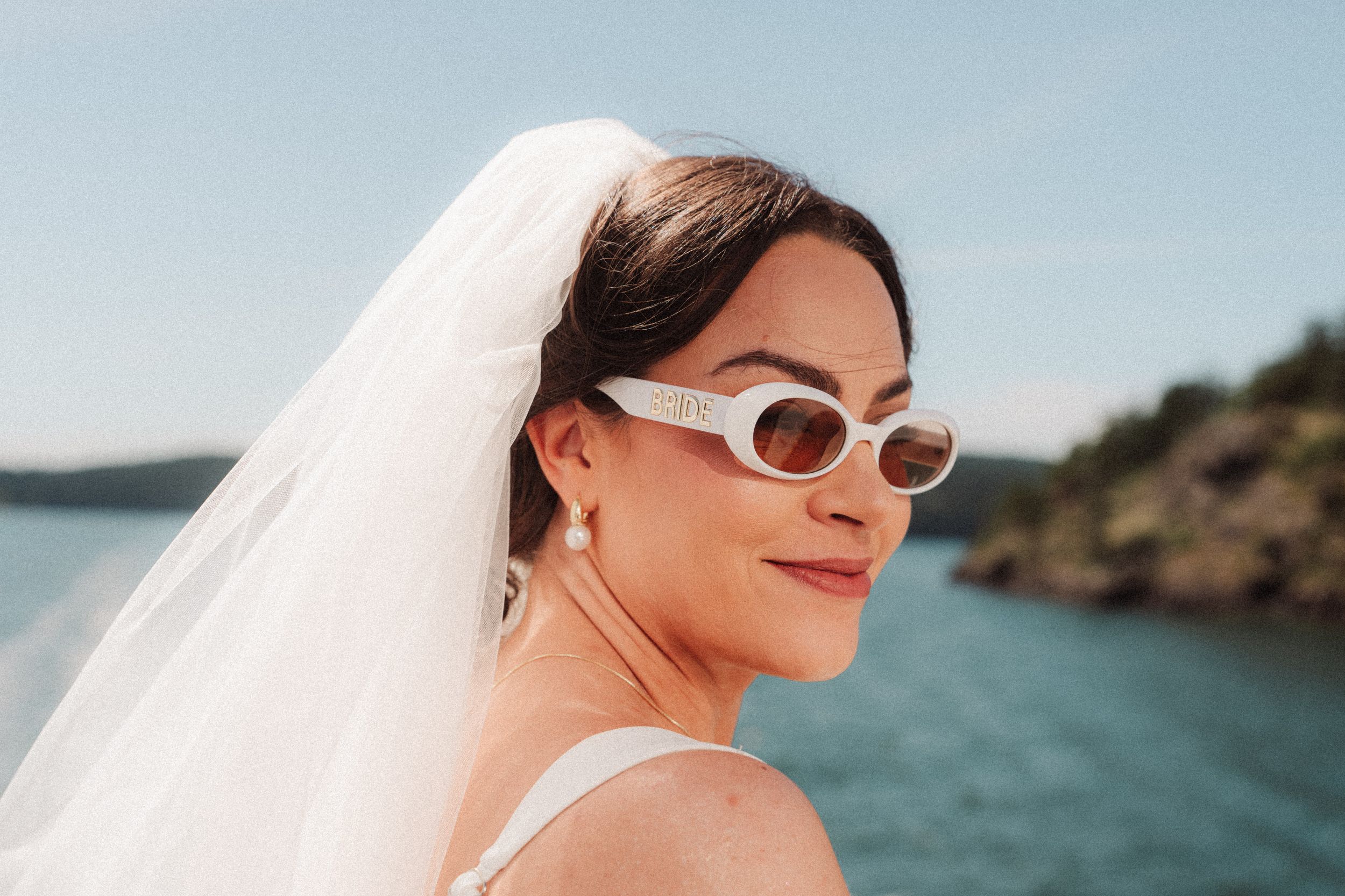 Smiling bride in white retro sunglasses and a flowing veil stands by calm blue water with a tree-covered shoreline behind her.