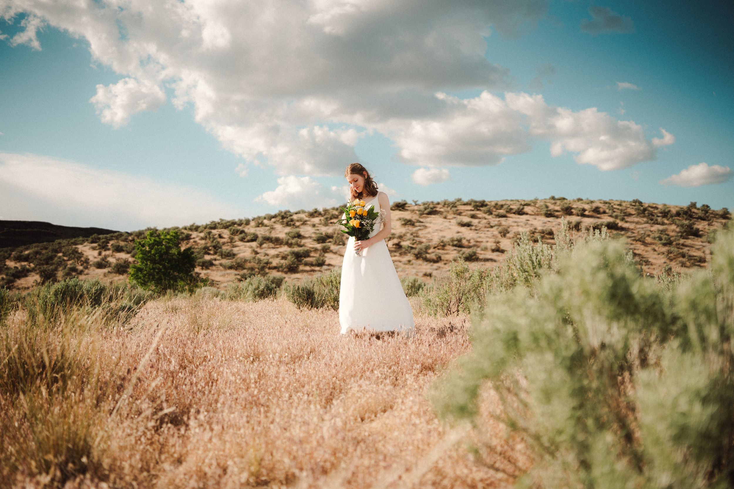 Bride in a white wedding dress holding a bouquet stands in a golden desert field with sagebrush under a bright blue sky and clouds.