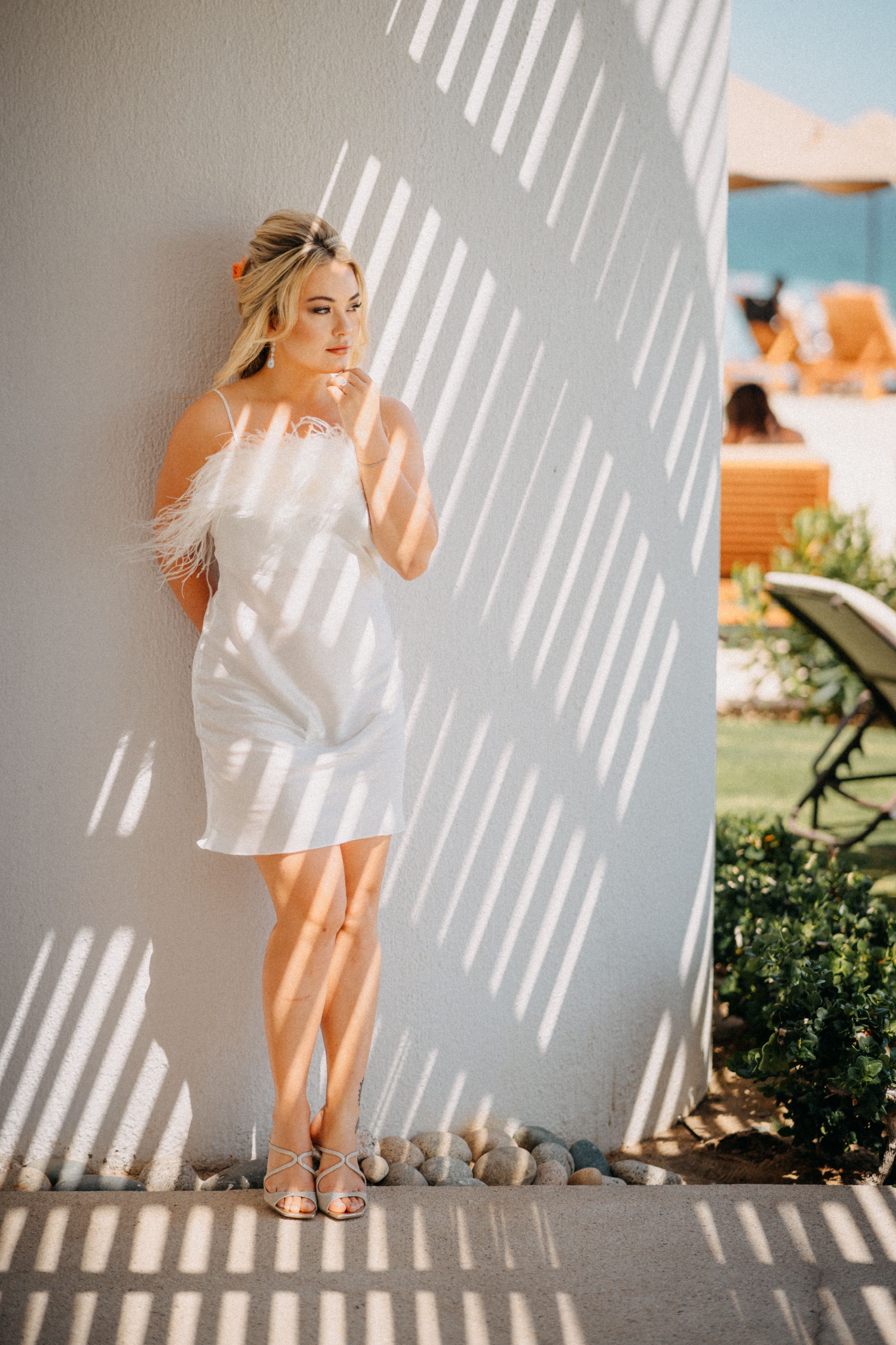 Woman in a white feathered mini dress and strappy sandals leans against a sunlit wall with striped shadows at a seaside resort.