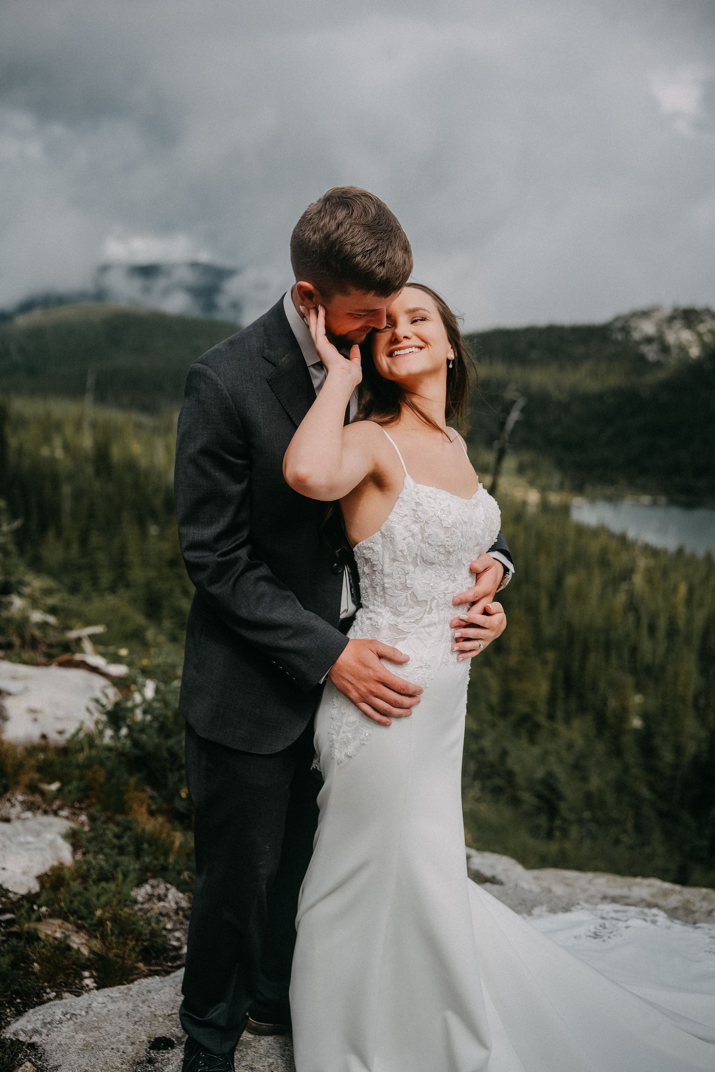 Bride in a white lace gown laughs as her groom in a dark suit embraces her on a rocky mountain overlook with forested valley and cloudy sky.