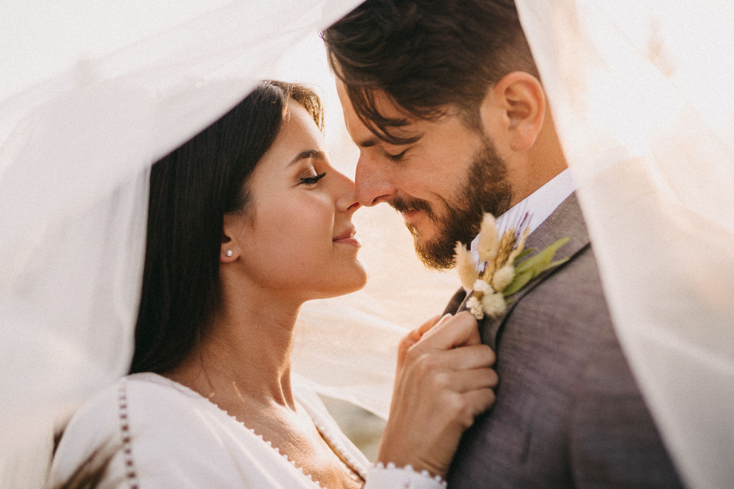 Smiling bride and groom touch noses under a sheer white veil, the groom wearing a gray suit with a floral boutonniere.