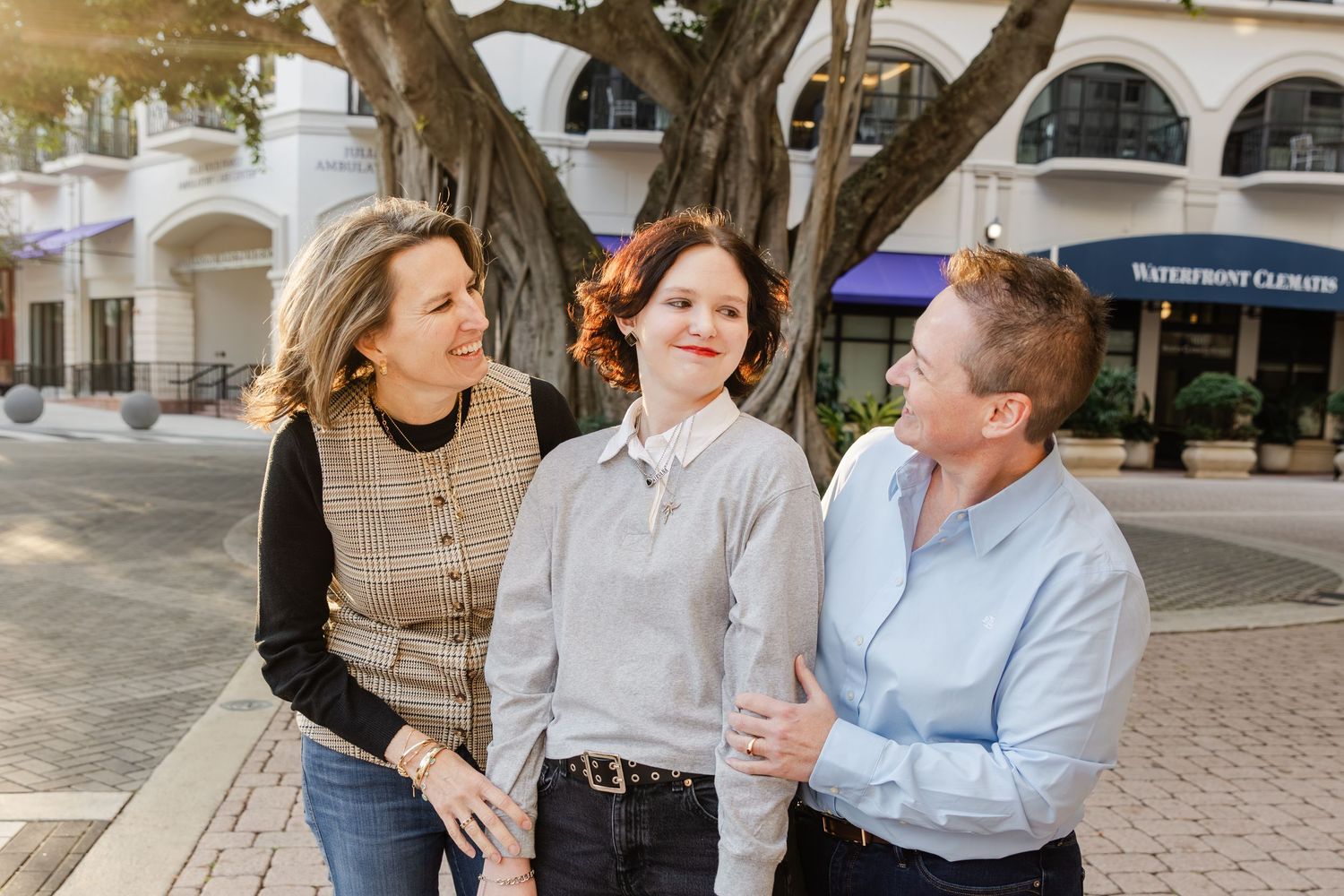 Three people smiling and talking outdoors near a large tree in a sunny waterfront shopping area downtown west palm beach.