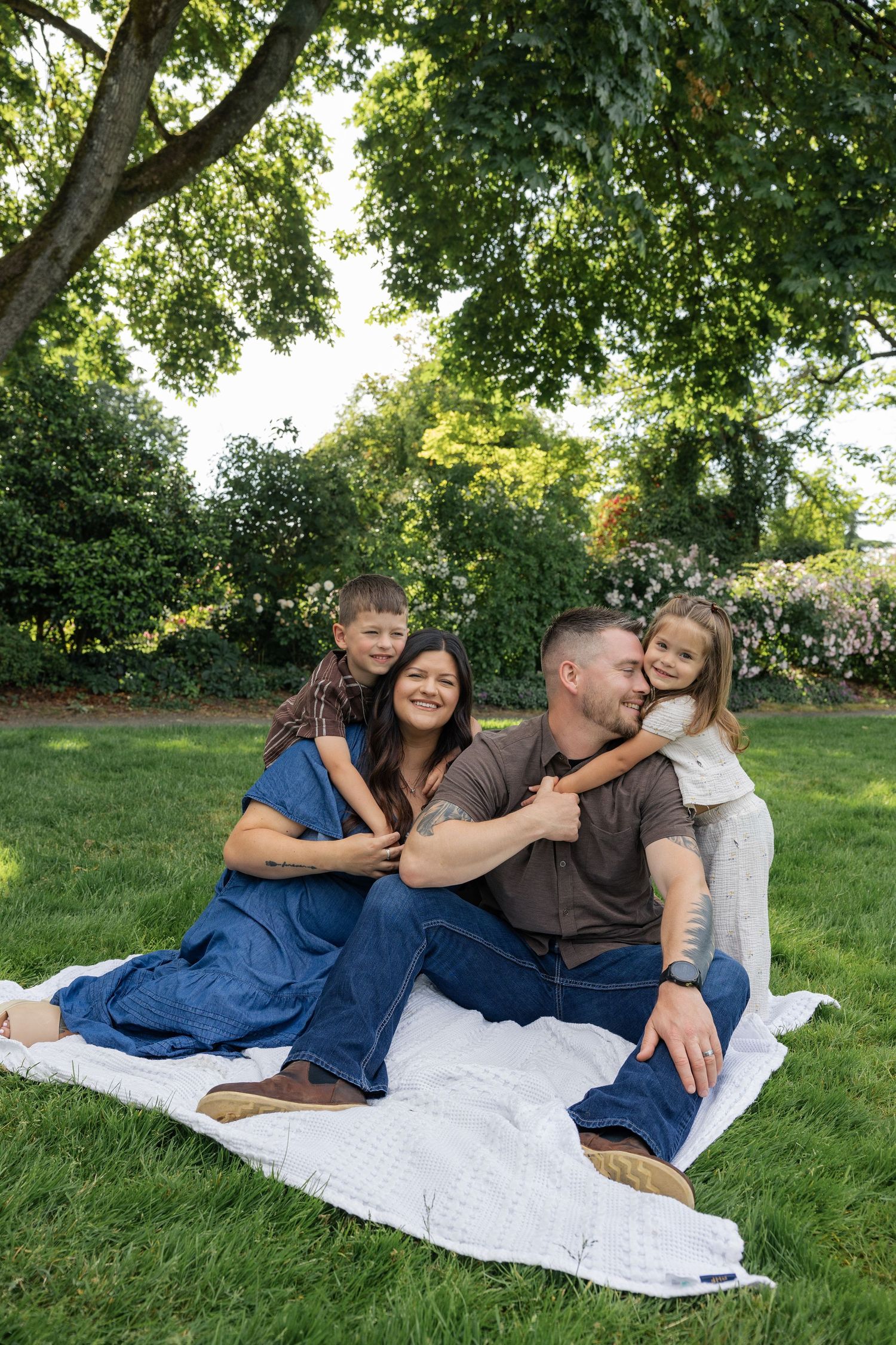 Happy family of four sitting on a blanket in a lush green park, parents smiling as children embrace them.