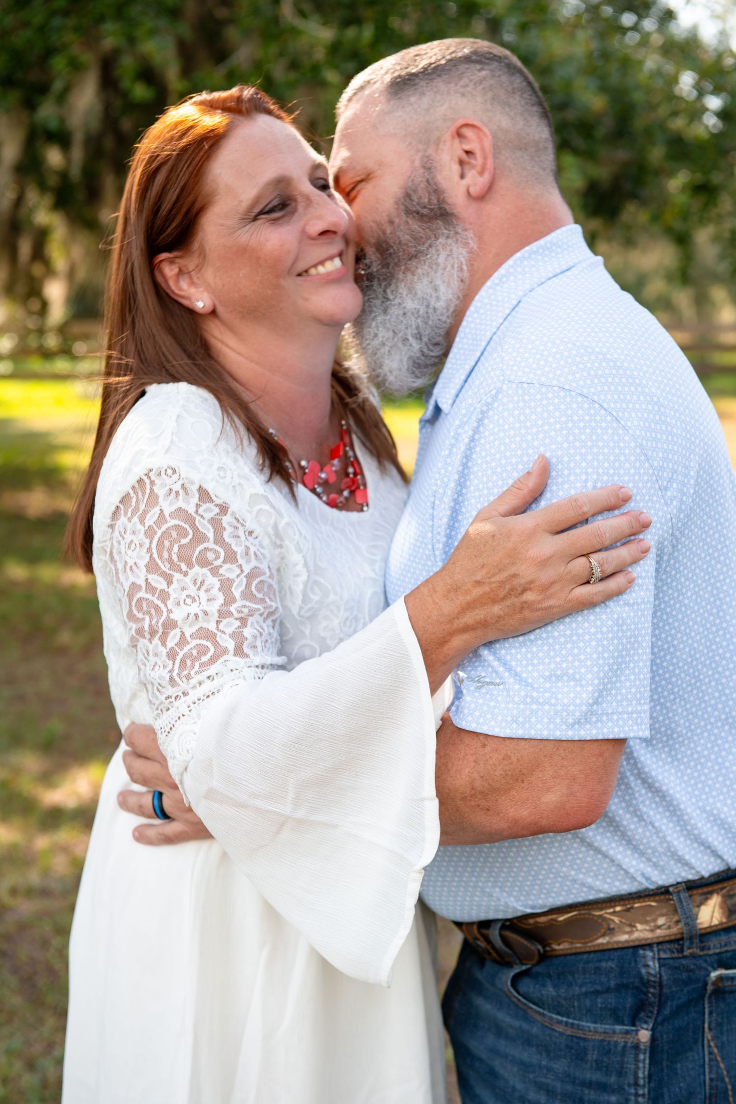 A couple shares a romantic moment outdoors, woman in white lace dress smiling as bearded man leans in for a kiss.
