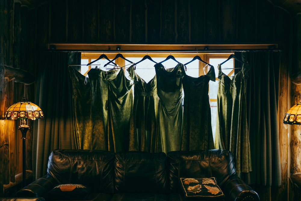 Dark bridesmaid dresses hanging on a rack above a leather couch in a dimly lit rustic room.