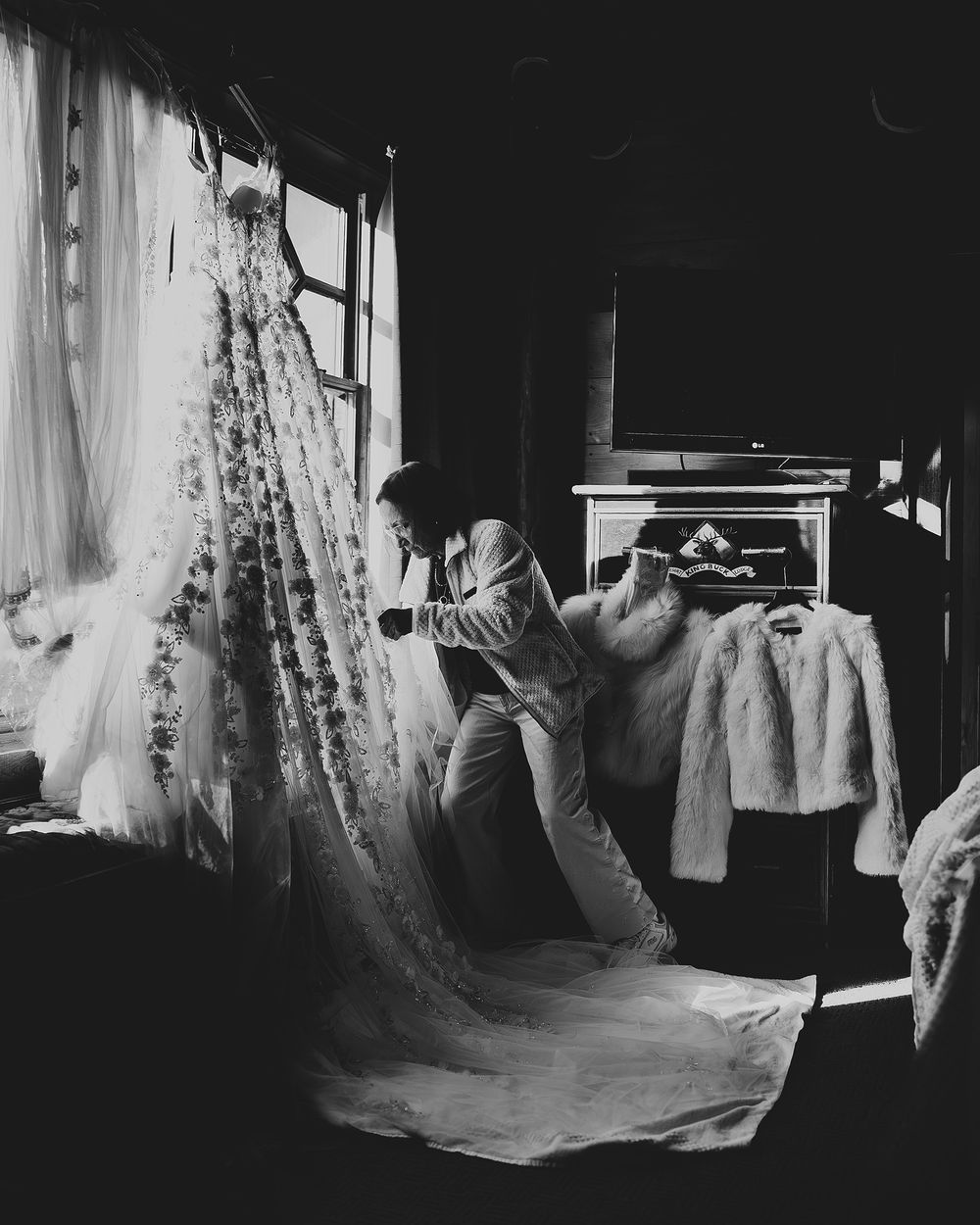 Black and white photo of a bride examining her wedding dress hanging by a window in a dimly lit room.