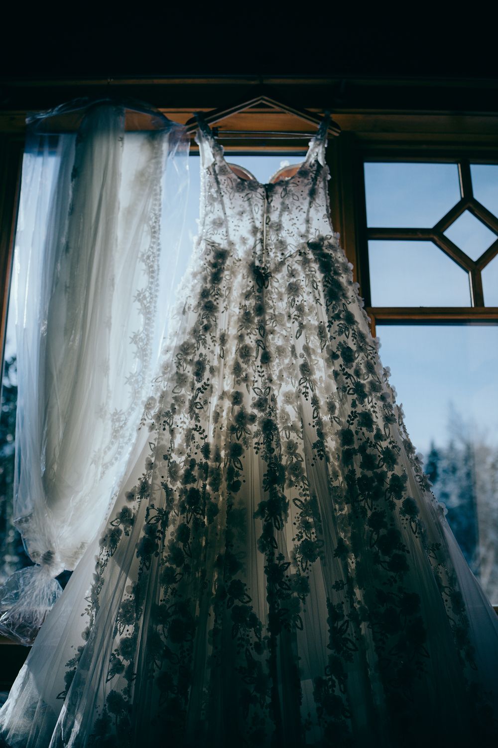 Elegant lace wedding dress with floral embroidery hanging by a window with sheer curtains and snowy trees outside.