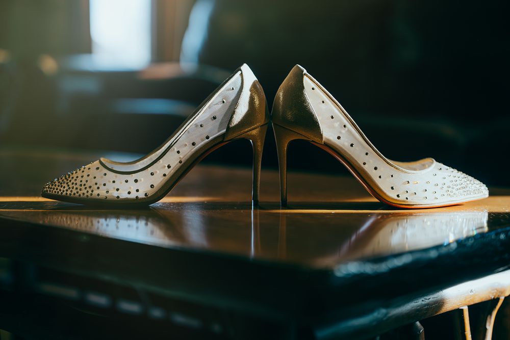 Elegant white high heel shoes with gold stilettos and rhinestone details displayed on a wooden surface.