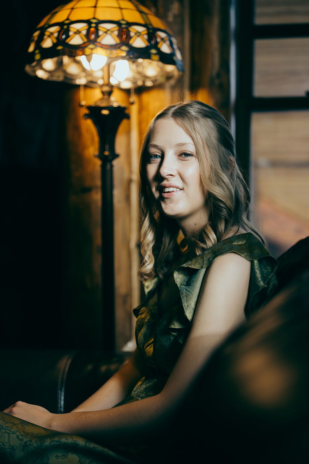 Young blonde woman smiling warmly in a dark room, seated near a glowing Tiffany-style floor lamp.