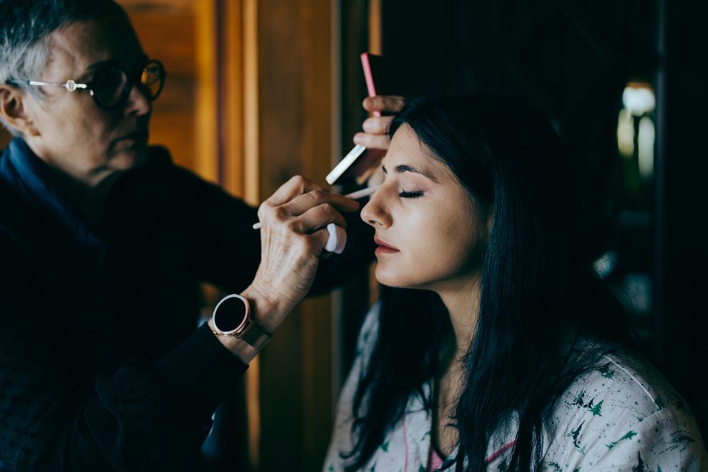A makeup artist carefully applies eyebrow makeup to a young woman with dark hair in a dimly lit room.