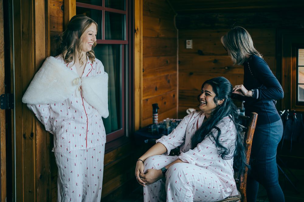Two women in floral robes getting ready in a rustic cabin, while a stylist works on one woman's hair.