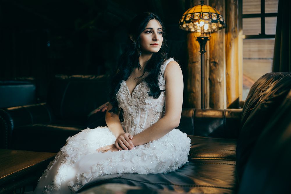 A young woman in a white ruffled dress sits gracefully on a dark sofa, lit by a vintage lamp in a moody interior.