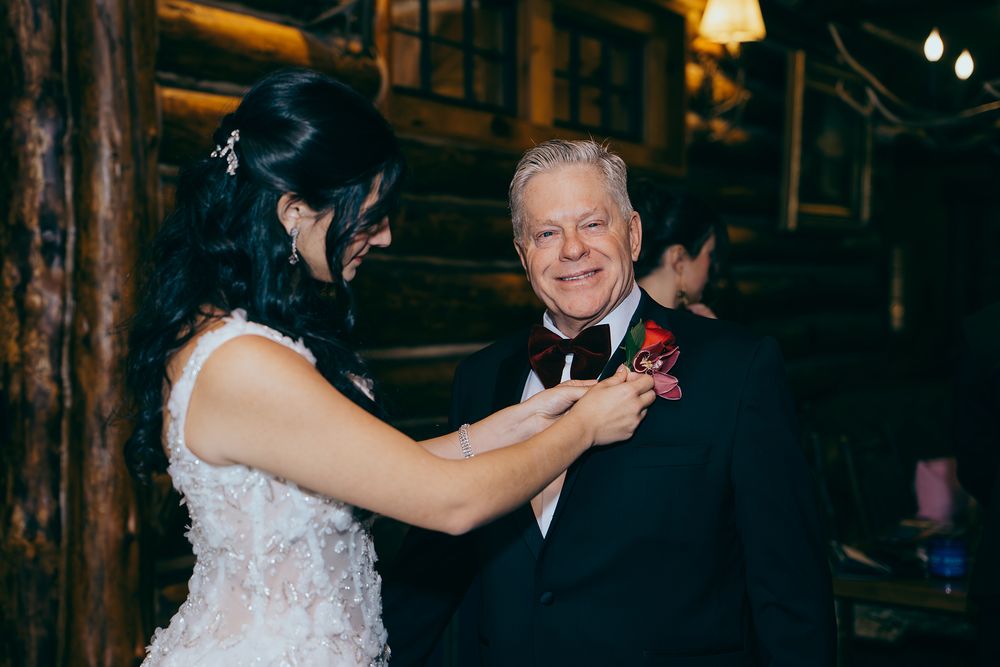 Bride pinning a boutonniere on an older man in a tuxedo at a rustic wedding venue.