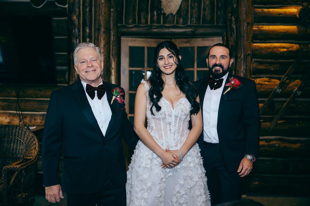 Bride in white lace gown poses with two men in tuxedos inside a rustic log cabin wedding venue.