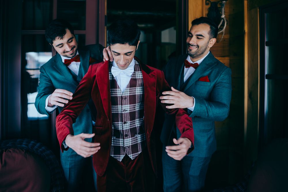 Groom in red velvet suit assisted by two groomsmen in gray tuxedos with red bow ties during wedding preparations.