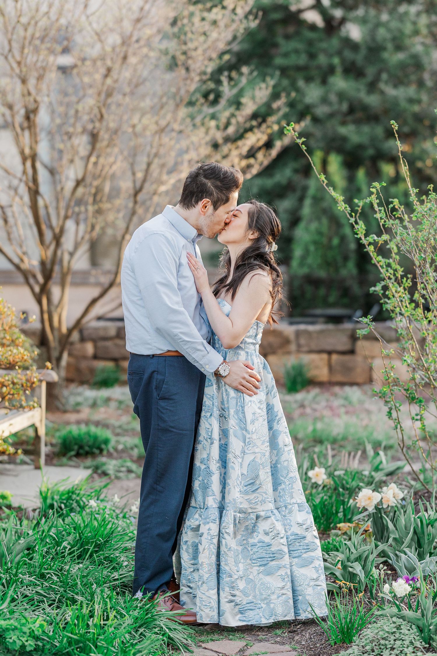 A couple shares a romantic kiss in a lush garden, the woman wearing a flowing blue floral gown during golden hour.