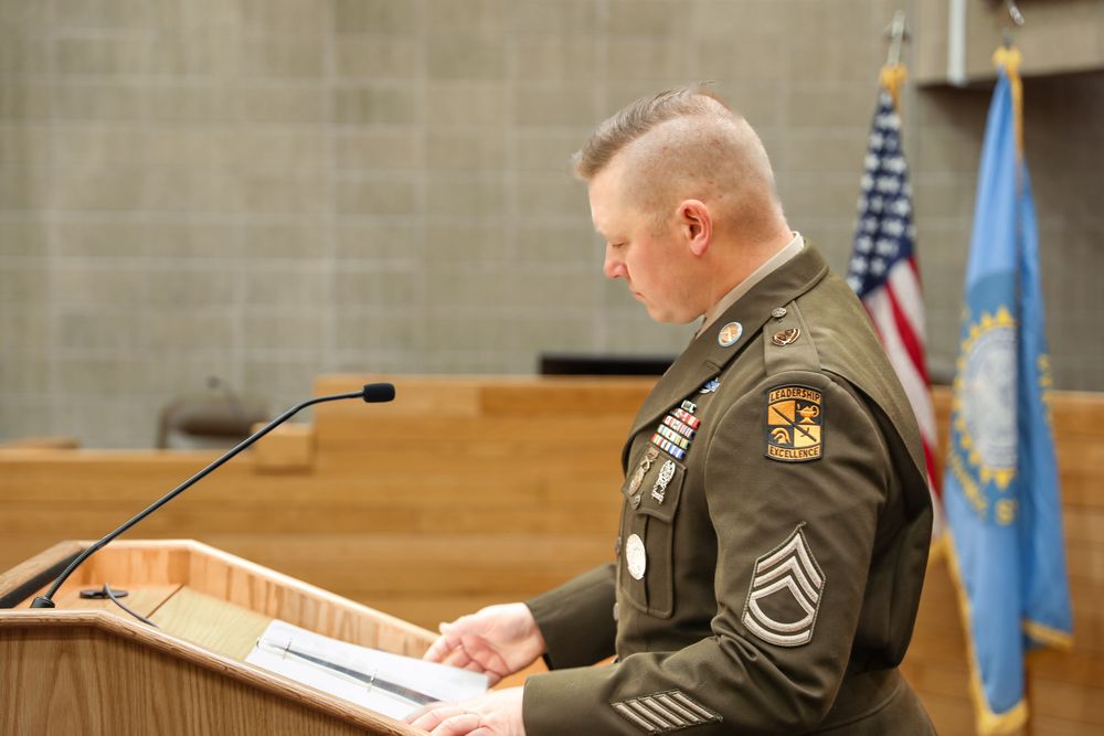 Military sergeant in dress uniform speaks at podium with American and state flags in background.