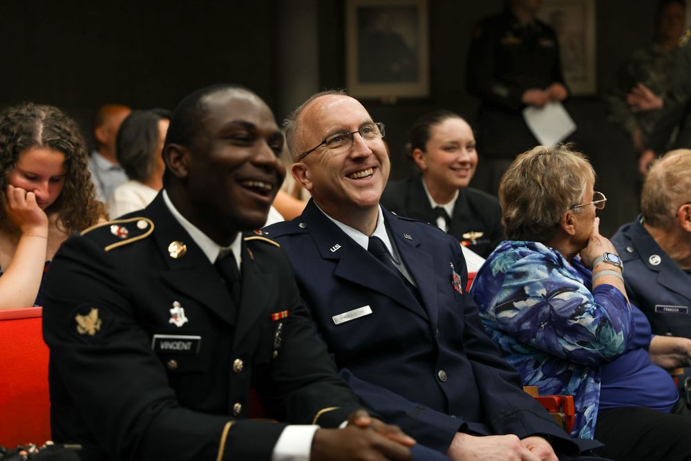 Two military officers in uniform smile and laugh together at a formal indoor event with other attendees in the background.