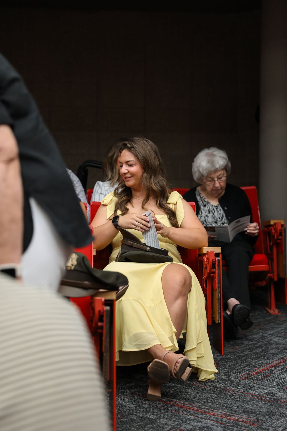 Woman in yellow dress seated in red auditorium chair, smiling, with elderly woman reading program in background.