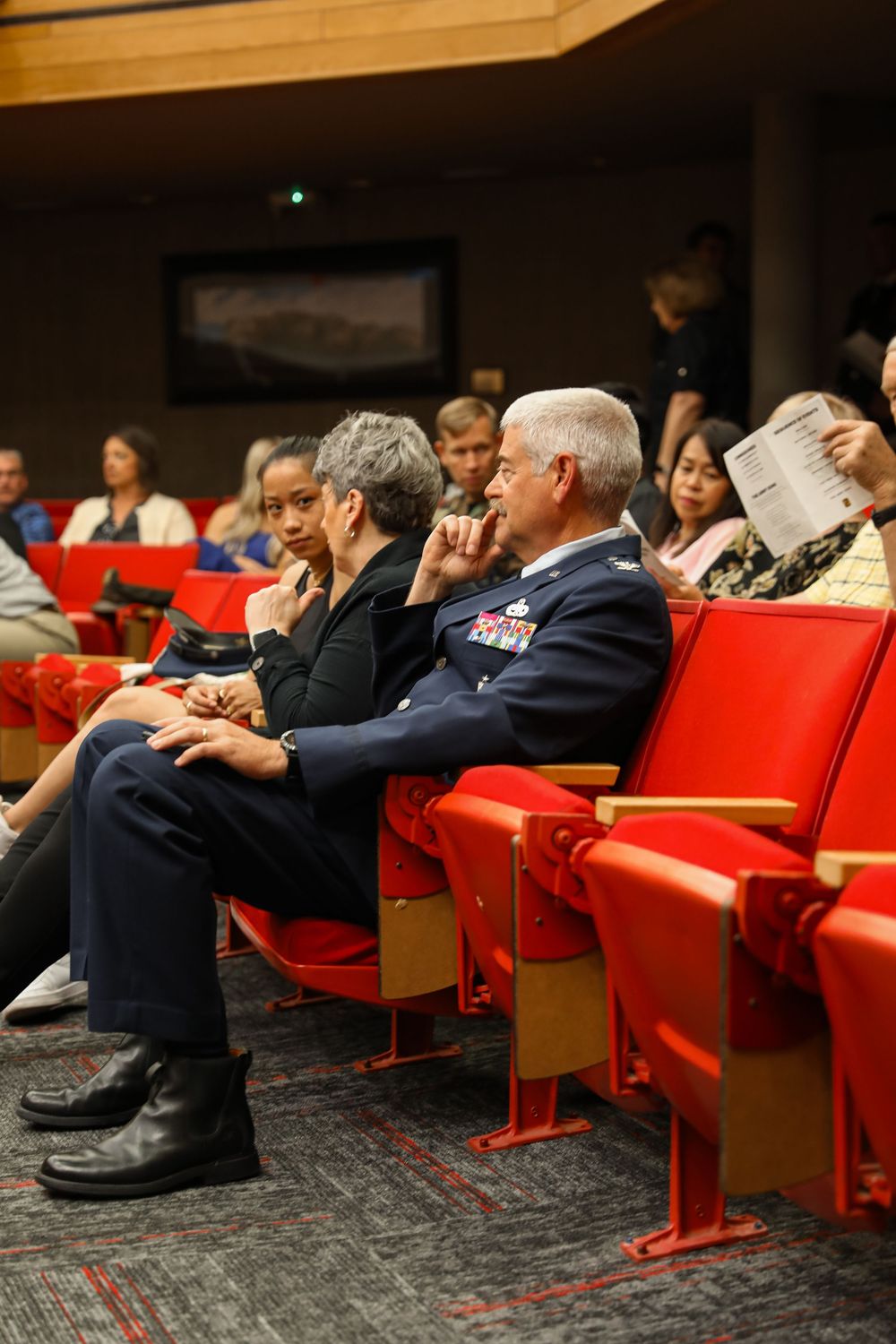 Military officer in dress uniform seated with civilians in red auditorium chairs at formal event.