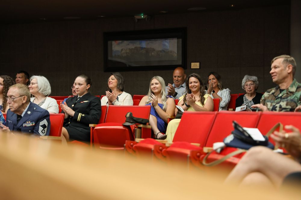 Audience of military personnel and civilians applauding in red-seated auditorium at formal ceremony.