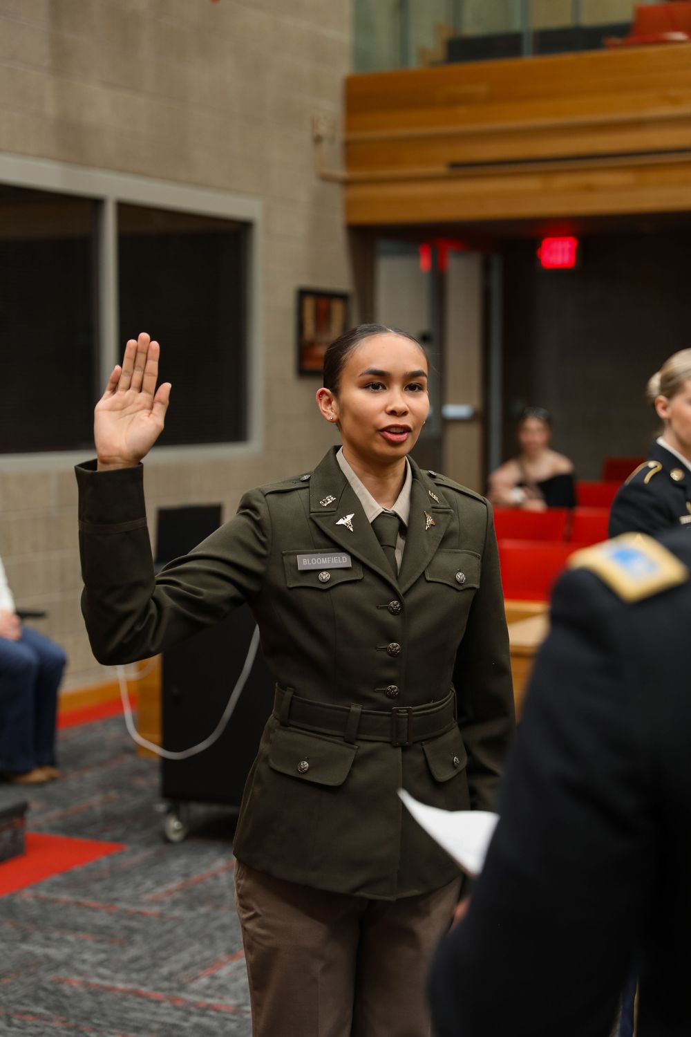 Female military officer in green dress uniform taking oath with raised right hand during formal ceremony indoors.