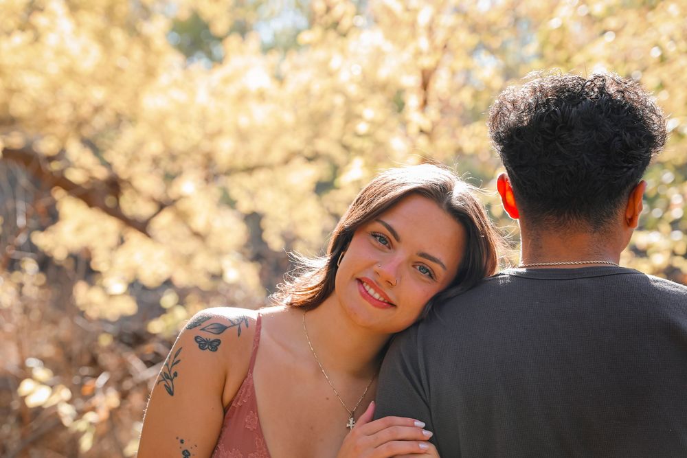 A smiling woman with a tattoo leans on a man's shoulder outdoors among golden sunlit trees.