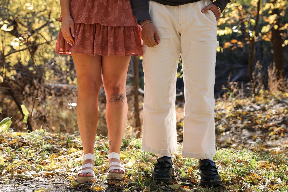 Couple standing outdoors in fall, woman in pink mini skirt, man in cream trousers and black shoes, autumn leaves on ground.