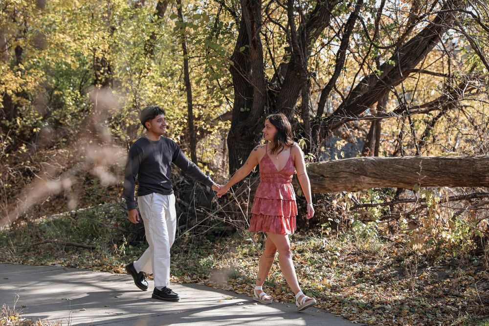Couple holding hands walking on a path through autumn trees in a park, man in gray and woman in pink dress.