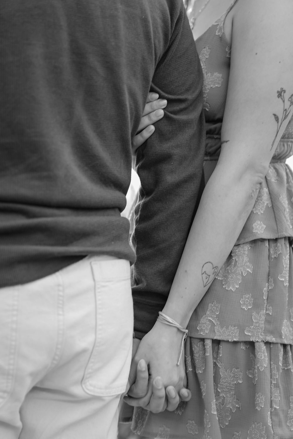 Black and white photo of a couple holding hands, woman with heart tattoo and bracelet wearing floral dress.