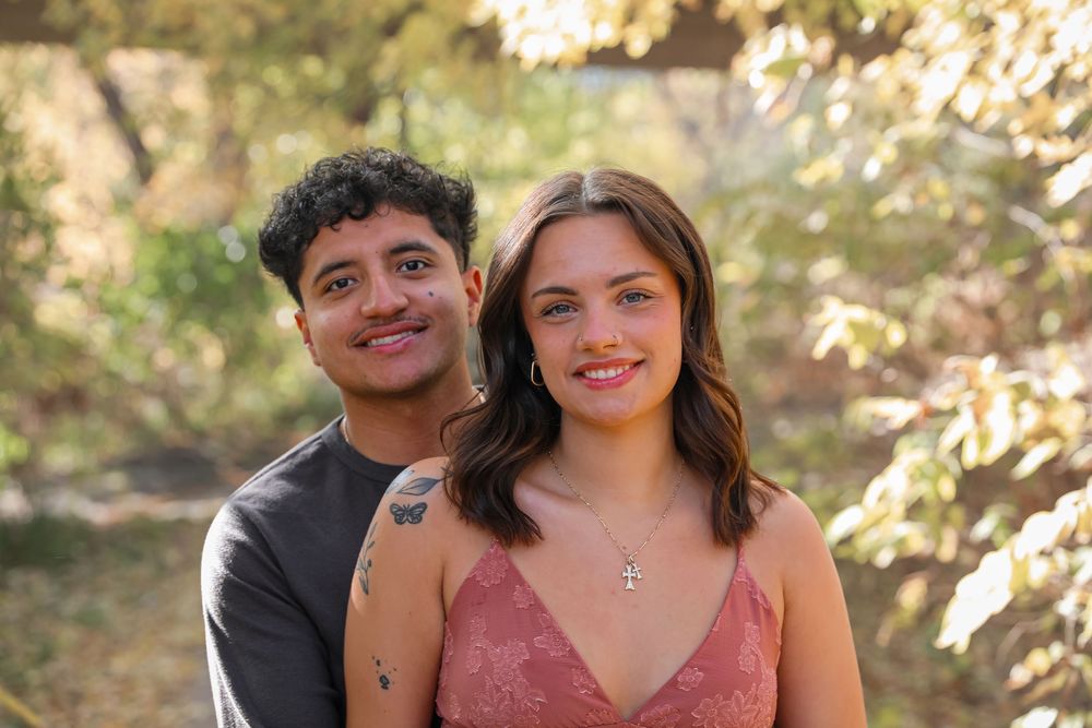 A smiling young couple poses outdoors among golden autumn trees, the woman wearing a pink lace top and cross necklace.