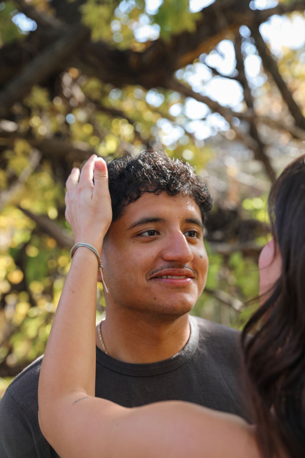 A young man smiles as a woman's hand gently touches his hair outdoors under a tree.