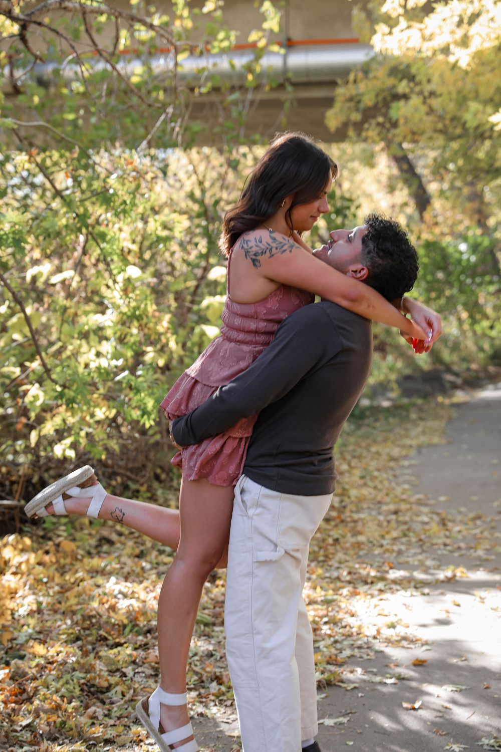 A man lifts a woman in a romantic embrace outdoors among autumn foliage on a scenic park path.