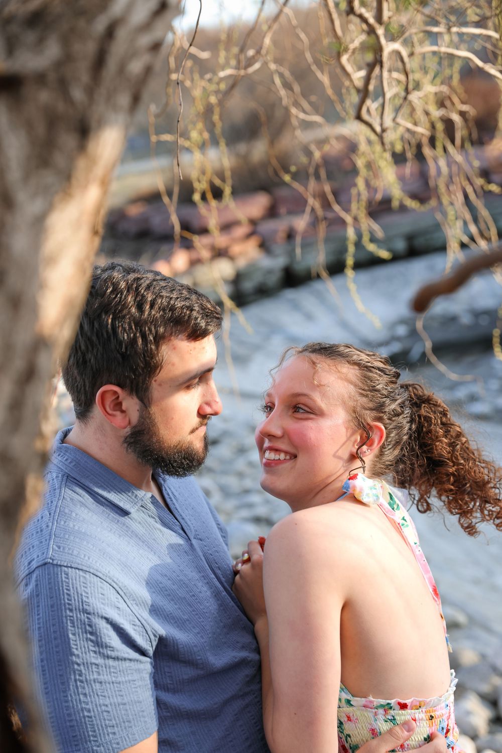 A smiling young couple sharing a romantic moment near a river, woman in floral dress and man in blue shirt.