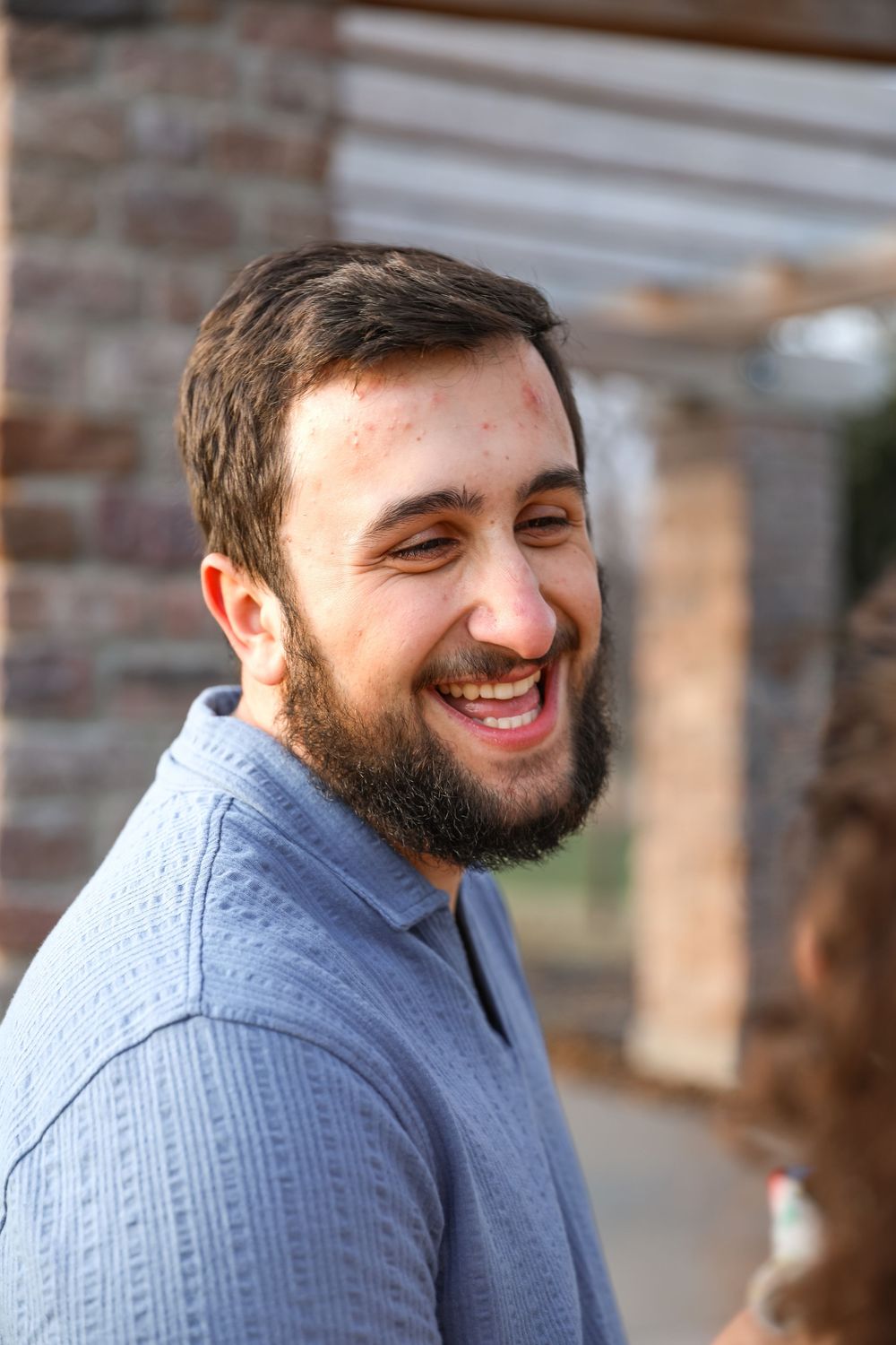 Smiling bearded man in blue polo shirt laughing outdoors near brick building.