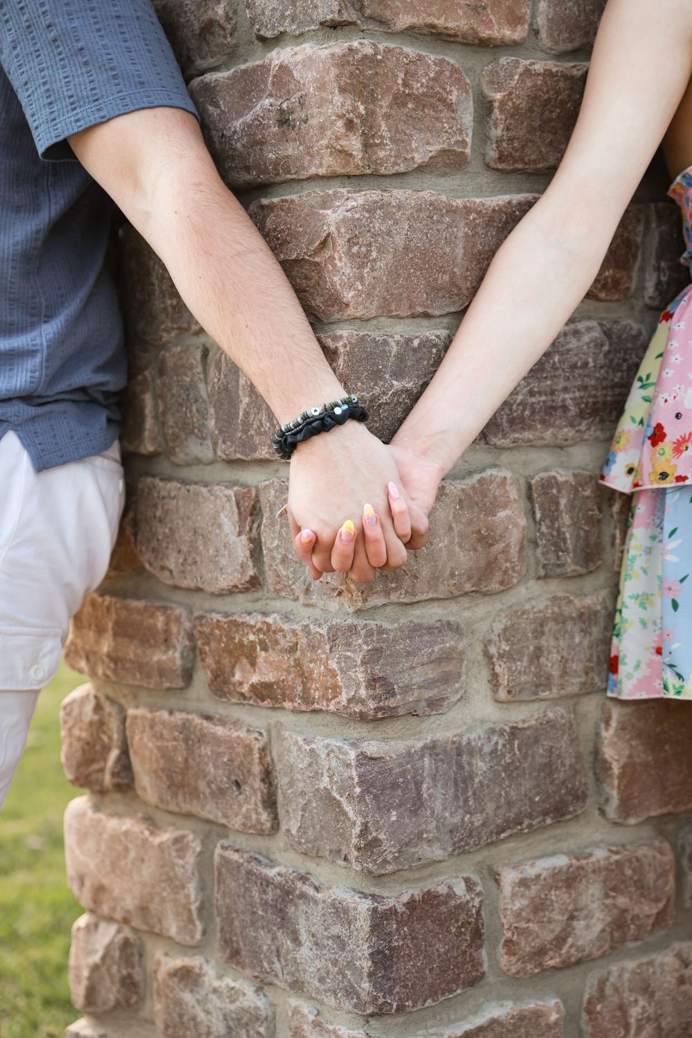 Couple holding hands behind a rustic brick pillar, one wearing a black beaded bracelet, outdoors.