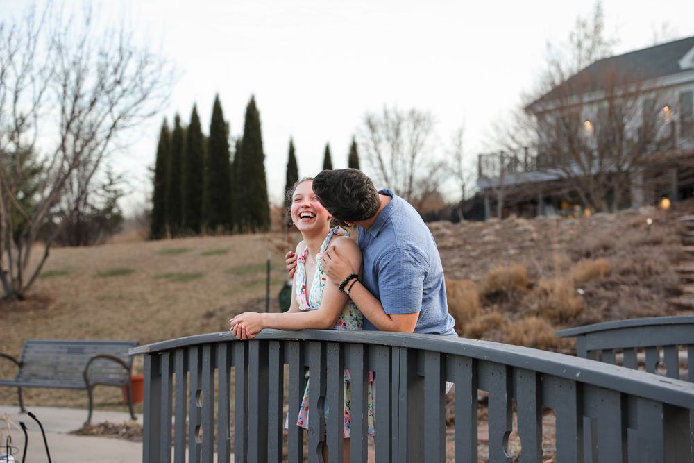 Happy couple sharing a romantic moment on a bridge outdoors, laughing and embracing in a park setting.
