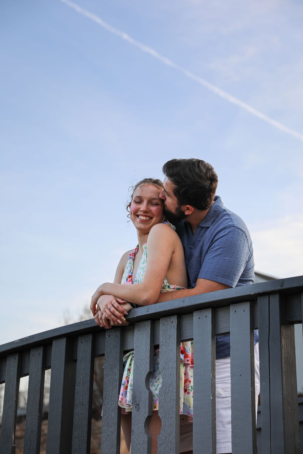 A smiling couple embraces on a wooden railing outdoors, man kissing woman's cheek against a blue sky.